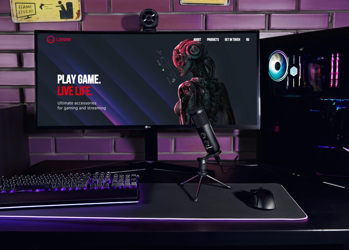 A LORGAR monitor showing the line Play Game. Live Life. on a setup with a microphone and backlit keyboard